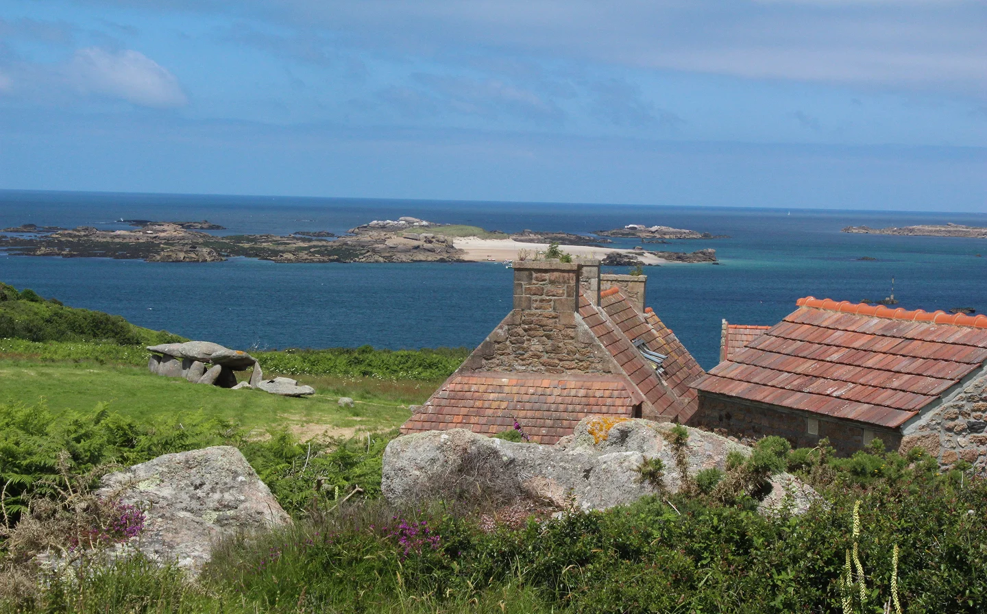 Vue depuis l'île Milliau sur la côte bretonne : au premier plan, des rochers granitiques et une végétation sauvage entourent les toits en tuiles rouges d'une maison en pierre, ainsi qu'un dolmen visible sur la gauche. En arrière-plan, la mer bleue s'étend jusqu'à un îlot rocheux avec une plage de sable blanc, sous un ciel partiellement nuageux.